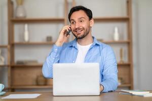 A man sitting at a desk, engaged in a phone conversation. He is multitasking, with a laptop open in front of him. photo