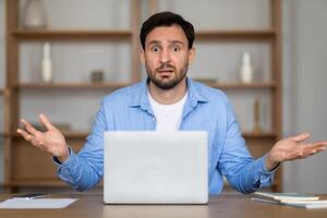 A man sits in a home office environment, displaying an expression of confusion as he looks at his laptop screen with his hands raised slightly in a questioning gesture photo