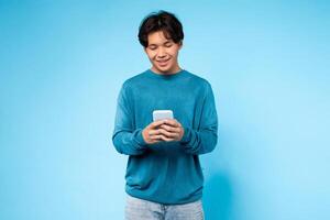 Asian guy wearing a blue sweater is sitting or standing and looking intently at a cell phone in his hands. He appears to be focused on the screen, possibly browsing, reading, or texting on the device. photo