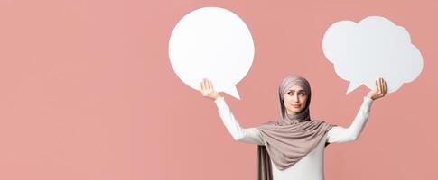 Difficult choice. Concerned arabian woman in hijab holding and choosing between two empty speech bubbles, pink studio background with free space photo