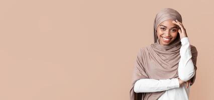 Shy modest black muslim girl in hijab touching her forehead and timidly looking at camera and smiling, yellow background with free space photo