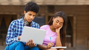 A teen black guy sits outside, using a laptop while a teen girl takes notes in a notebook. They appear to be engaged in a learning or studying activity, perhaps a school project photo