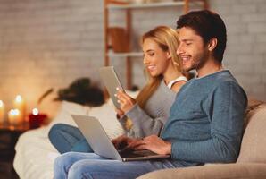 Gadget Users. Smiling Couple Using Laptop And Tablet Computers Sitting On Couch Indoor. Selective Focus photo