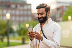 Indian man with a beard is smiling and looking at his smartphone while standing in an urban park during sunset. He wears a white shirt and carries a shoulder bag photo