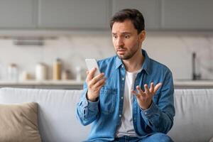 A man sits on a light gray couch in a kitchen, staring at his smartphone with a confused expression. He has his right hand raised, gesturing with his fingers, while holding the phone in his left hand. photo
