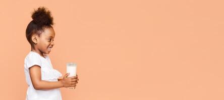 A little back girl with curly hair smiles as she holds a glass of milk in front of a peach-colored background. She is wearing a white shirt with ruffles and looks happy and healthy, copy space photo