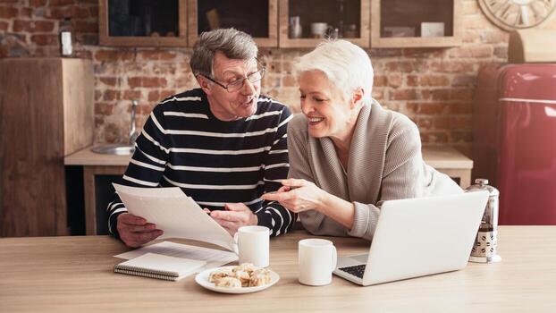 un más viejo Pareja se sienta a un cocina mesa, revisando documentos mientras disfrutando un taza de café. el hombre es participación un papel en su manos mientras el mujer puntos a alguna cosa en el papel. foto