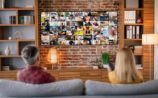 A couple sits on a couch in a living room, facing a large television screen displaying a mosaic of images. The room features a brick wall and wood accents, suggesting a comfortable and cozy atmosphere photo