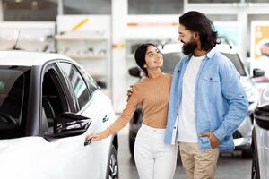 Indian couple standing next to a car in a parking lot. They are both looking towards the camera, with the car door open and the trunk slightly ajar. The man is holding car keys in his hand. photo