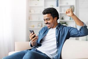 African American man is seated on a couch indoors, holding a cell phone in his hand. He appears focused on the device, with a neutral expression on his face, gesturing photo