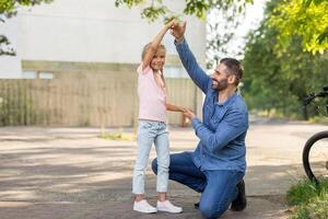 A father kneels down to play with his daughter on a sunny day. They are both smiling and having fun outside. There is a bicycle in the background. photo