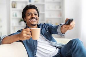 A cheerful African American man is lounging comfortably on a sofa with a warm beverage in one hand and a TV remote in the other, seemingly in the middle of selecting a program to watch photo
