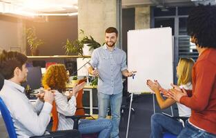 A man stands in front of a group of attentive individuals, gesturing and speaking confidently as he delivers a presentation. The audience is seated, listening intently, taking notes photo