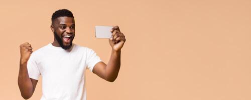 African American man dressed in a white t-shirt, with a beard, and a huge smile. He is holding a smartphone and taking a selfie. He is clearly excited and appears to be celebrating a win, copy space photo
