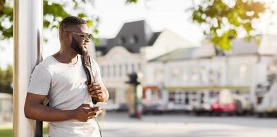 A man in sunglasses uses his phone while leaning against a pole in a city setting. photo