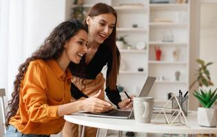 Two Happy Ladies Using Laptop Computer Working And Learning Online Together Or Making Call Sitting At Desk In Modern Office. E-Learning. Females Entrepreneurship Career Concept. Side View photo