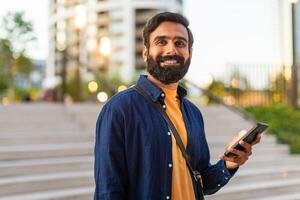 Indian man stands in an urban park, smiling as he holds his smartphone. He appears cheerful and relaxed in a casual outfit, while modern buildings and greenery surround him. photo
