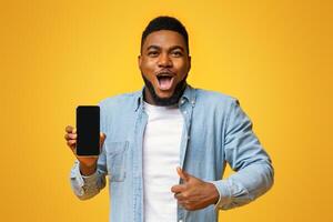 Wow, amazing app. Surprised african man holding smartphone with black screen and showing thumb up, yellow background with free space photo