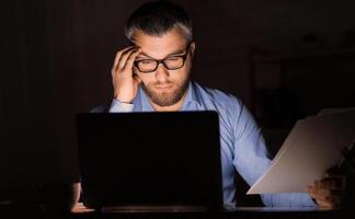 A man in glasses is seen working late in a dimly lit office. He holds documents in one hand while looking intently at a laptop screen. The setting suggests diligent effort and focus photo