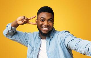 Say Cheese. Joyful black millennial guy showing peace sign while taking selfie, yellow studio background with free space photo