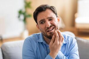 A man in discomfort is seated on a couch, holding his face in pain due to a toothache. His expression reveals his suffering as he tries to alleviate the ache. photo