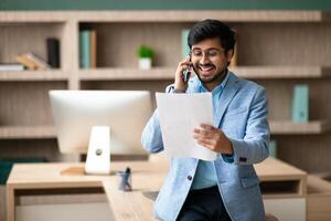 A smiling Indian man in a blue blazer is talking on the phone while reading documents. He is standing at a desk in a modern office. photo
