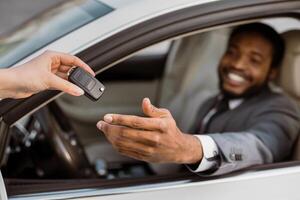 African American man is sitting in the drivers seat of a car with a person handing him the keys. He is smiling and reaching out to take them. photo