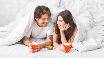 A couple, a man and a woman, are lying in bed, covered by white bedding. They are both smiling and looking at each other, holding mugs of coffee and a plate of cookies. photo