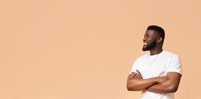 A smiling black man stands against a peach background with his arms crossed. He is wearing a white t-shirt and looking off to the side. The man is confident and relaxed, copy space photo