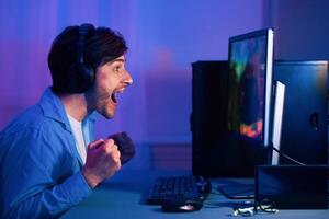 A young man wearing headphones sits at his computer, excitedly playing a game. His mouth is open in surprise and his fists are clenched in victory as he watches the action unfold on the screen. photo