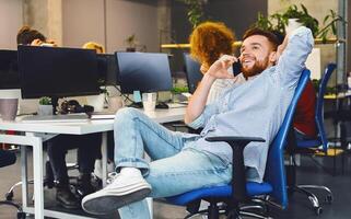 A man in business attire is sitting in an office chair, engaged in a phone conversation on his cell phone. The setting is a typical office environment with a desk and computer in the background. photo