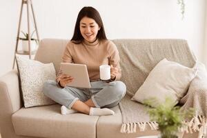 A woman relaxes on a sofa while holding a tablet and a cup of coffee. She is dressed in casual clothing and has a relaxed expression on her face. photo