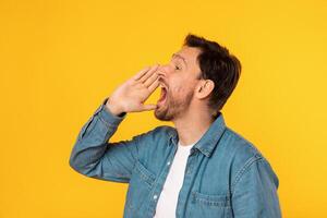 Young man stands against vivid yellow backdrop, his mouth wide open as if shouting or calling out loudly. He is casually dressed in denim shirt and appears to be making announcement with fervor. photo