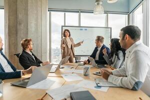 A businesswoman stands in front of a large screen and points to a graph as she presents data to a team of colleagues seated around a table in a modern office. photo
