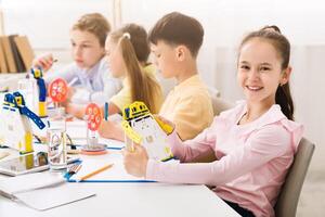This image shows a group of children in a classroom setting, engaged in a STEM activity. They are working on building robots, and the girl in the foreground is holding up her finished creation photo