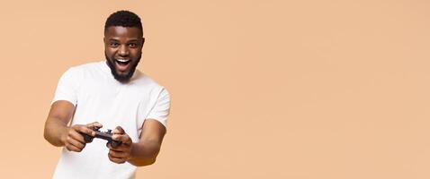 A young African American man wearing a white T-shirt is smiling excitedly as he plays games. He is holding a black controller in both hands, pointing it towards the camera, copy space photo