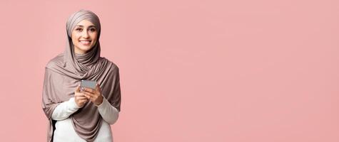 Best Apps for Muslims Concept. Smiling islamic girl in headscarf holding smartphone and looking at camera over pink studio background, free space photo