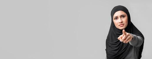 It's your fault. Concerned muslim woman in hijab pointing finger at camera, blaming somebody, posing over grey background with empty space photo