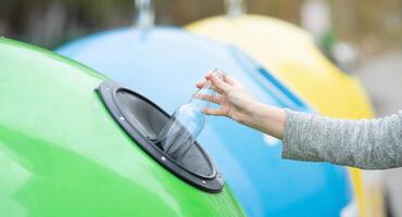 Waste Sorting And Recycling Concept. Unrecognizable female throwing empty glass bottle into green recycle bin garbage container outdoors, caring about environment, cropped image with selective focus photo