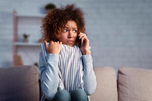 Scared afro woman calling phone and embracing pillow on sofa, afraid of noise near house, free space photo