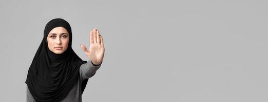 No abuse and discrimination. Muslim woman in hijab making stop gesture with her hand, covering her face with open palm over gray background photo