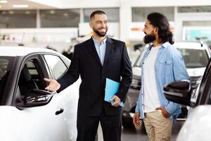 A smartly dressed car salesman, with a friendly demeanor, stands next to a shiny new car in a well-lit vehicle showroom, engaging with a potential customer Indian guy who is holding a brochure. photo