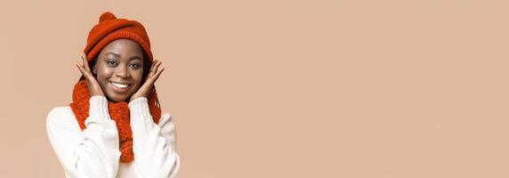 Portrait of sweet african woman in winter red hat touching her cheeks with hands over beige background, panorama with copy space photo