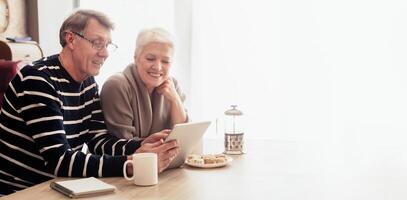 A happy senior couple sits at a table in their home, browsing the internet on a tablet. The man is wearing a striped sweater and glasses, while the woman is wearing a beige sweater and smiling. photo