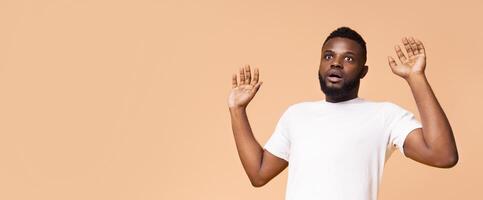 A Black man with a beard is wearing a plain white t-shirt and is standing in front of a beige background with his hands raised in front of him as if in surprise or alarm, copy space photo
