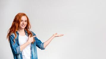 A cheerful redhead woman wearing a denim shirt over a white top is standing against a plain light background. She is looking at the camera with a smile while making a presenting gesture, copy space photo