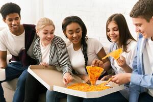 A group of friends sitting on a couch, enjoying slices of pizza. The diverse group is engaging in casual conversation while savoring their meal together. photo