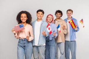 Multiethnic group of young adults stand together holding flags of different countries. They are all smiling and looking at the camera. The background is a simple white wall. photo