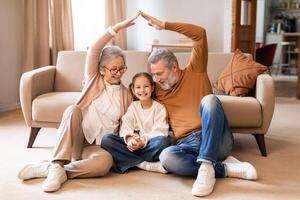 A joyful elderly couple sits on the floor with their young granddaughter between them, smiling and creating roof shape with their hands over the girls head photo