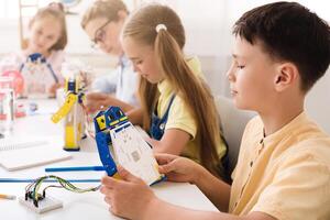 A young boy sits at a table in a classroom and builds a robot with other children. The boy is concentrating on his work as he holds a piece of the robot and examines it. photo